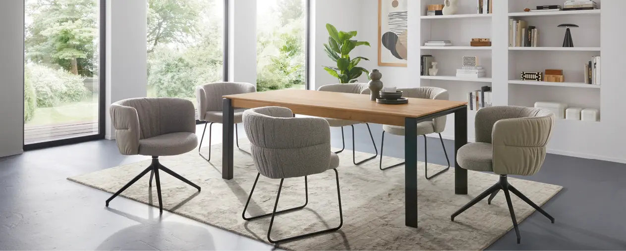 Helles Esszimmer mit massivem Holztisch, komfortablen gepolsterten Esszimmerstühlen und modernem Teppich. Große Fenster sorgen für viel Tageslicht und schaffen eine wohnliche Atmosphäre im modernen Wohnstil.